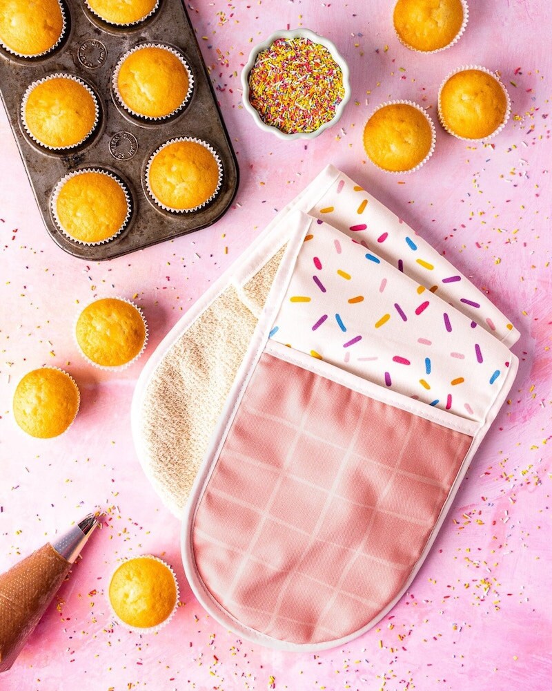 A double oven glove with a rainbow sprinkles pattern surrounded by baked cupcakes.