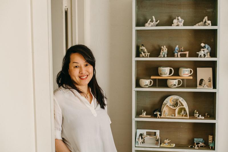 Artist Nicky Sa-eun Schildkraut next to shelves with a selection of her handmade ceramic mugs and mini sculptures.