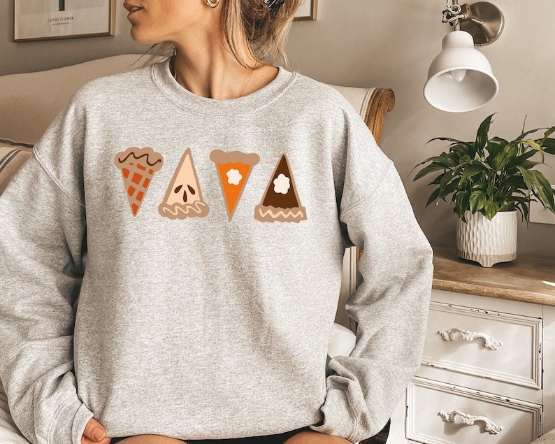 A woman wearing a gray sweatshirt featuring slices of different Thanksgiving pies on it.