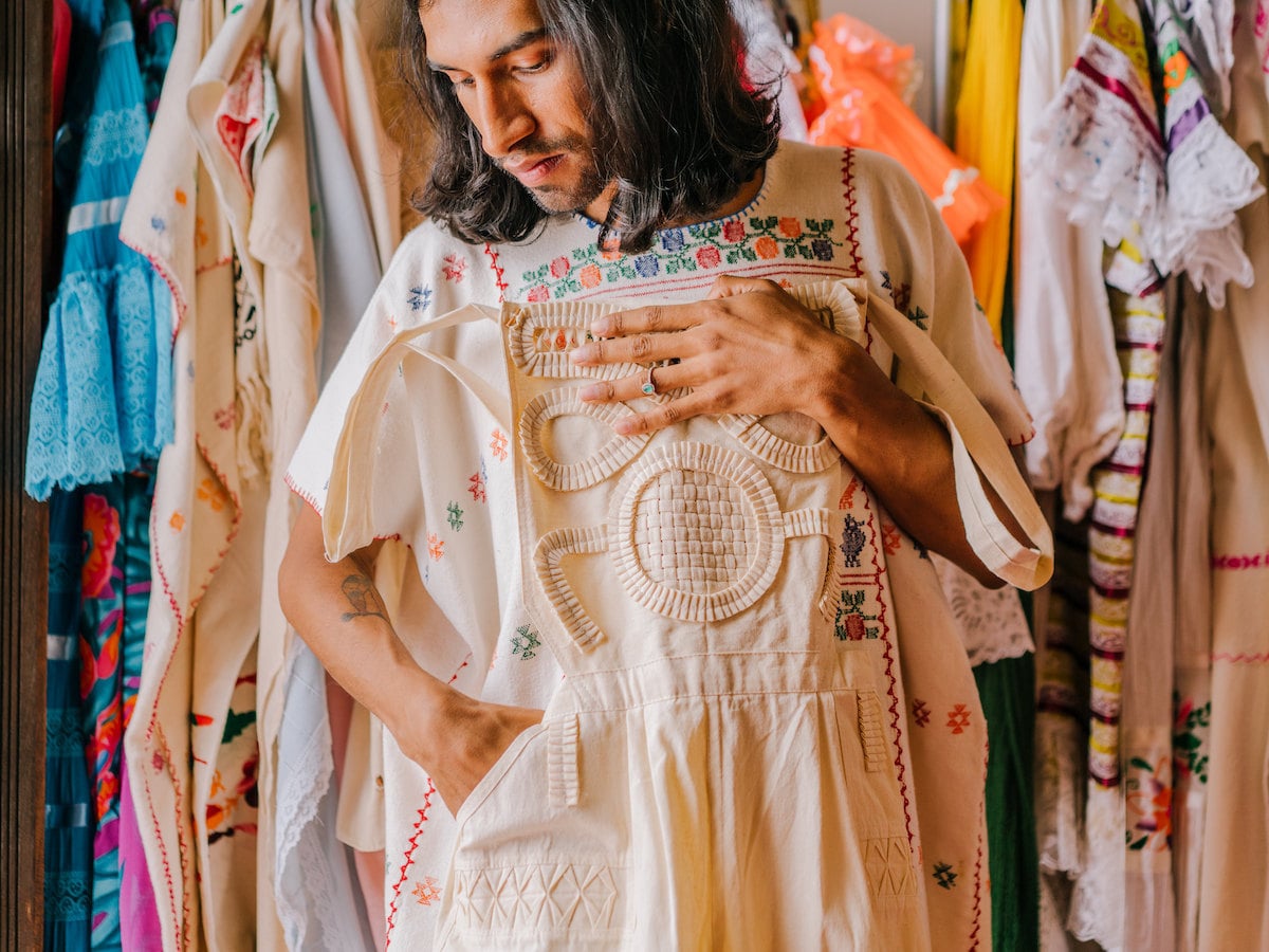 Jesus Herrera stands in front of a rack of clothes, and holds up an intricately embroidered Mexican garment. Jesus Herrera stands in front of a rack of clothes, and holds up an intricately embroidered Mexican garment.