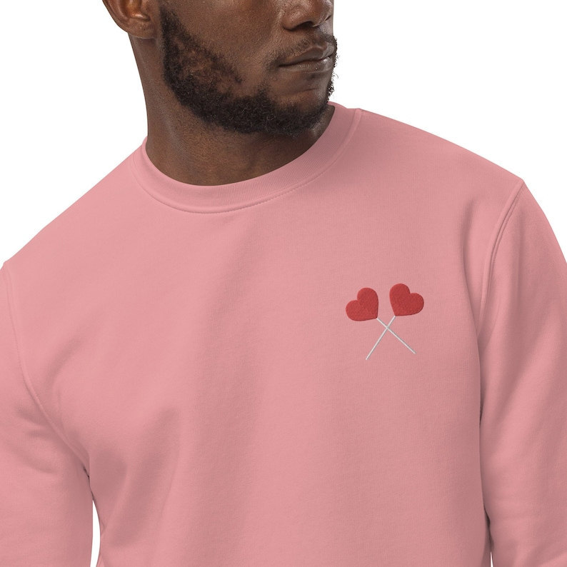 A Black man wearing a pink sweatshirt featuring two heart lollipops embroidered on it.