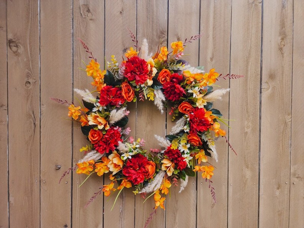 Fall wreath styles 2023: A bright fall wreath featuring red and orange flowers against a wood background.