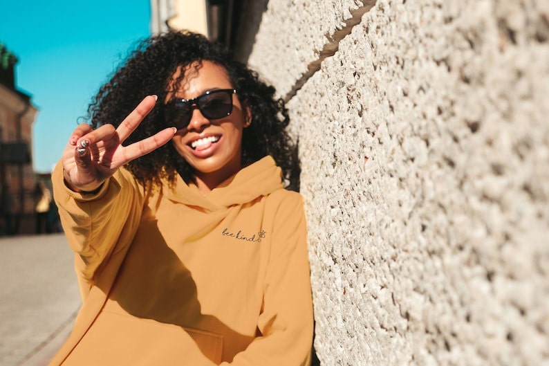 A Black woman smiling and giving the peace sign while wearing a mustard yellow sweatshirt with