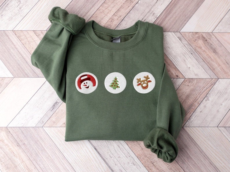 A folded green sweatshirt with holiday sugar cookie designs on it featuring a snowman, Christmas tree, and a reindeer.