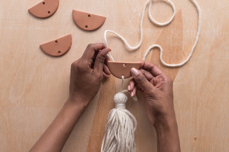 Image of hand pulling yarn through the bottom hole of a clay semicircle. Image of hand pulling yarn through the bottom hole of clay semicircle.