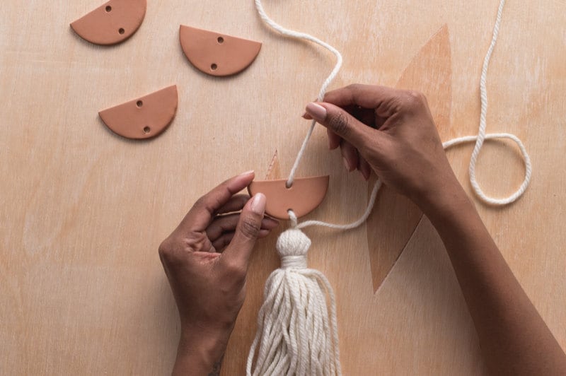Image of a hand pulling yarn through the top hole of a clay semicircle. Image of a hand pulling yarn through the top hole of a clay semicircle.