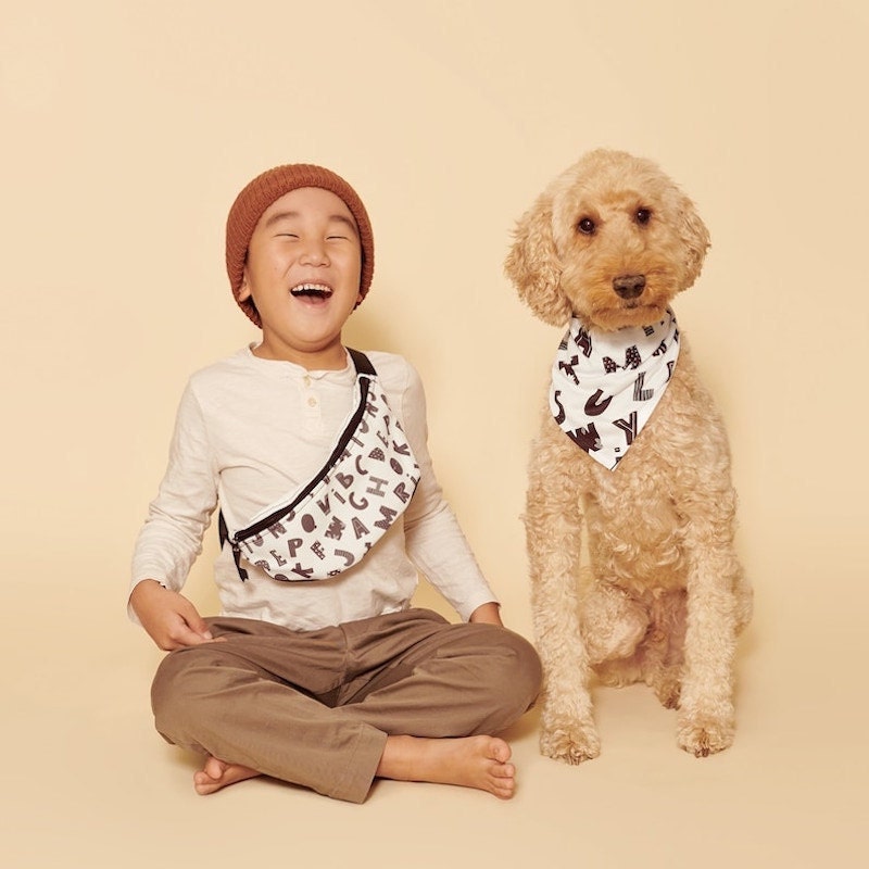 A little boy wears an alphabet-printed fanny pack sitting next to a fluffy golden dog who wears a matching bandana