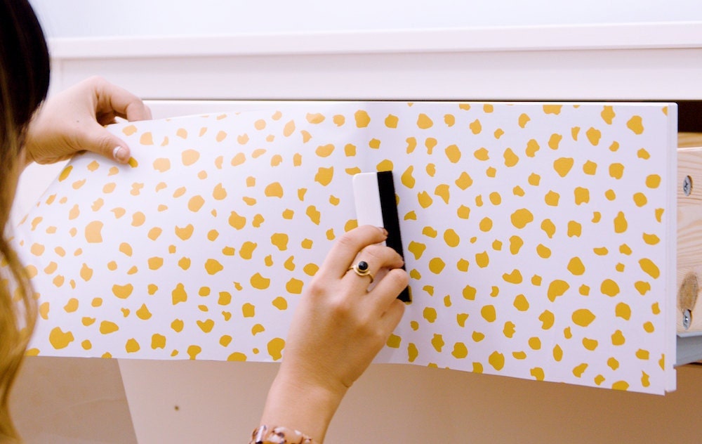 A woman smooths out air bubbles with a smoothing tool as she applies peel-and-stick paper to a dresser drawer