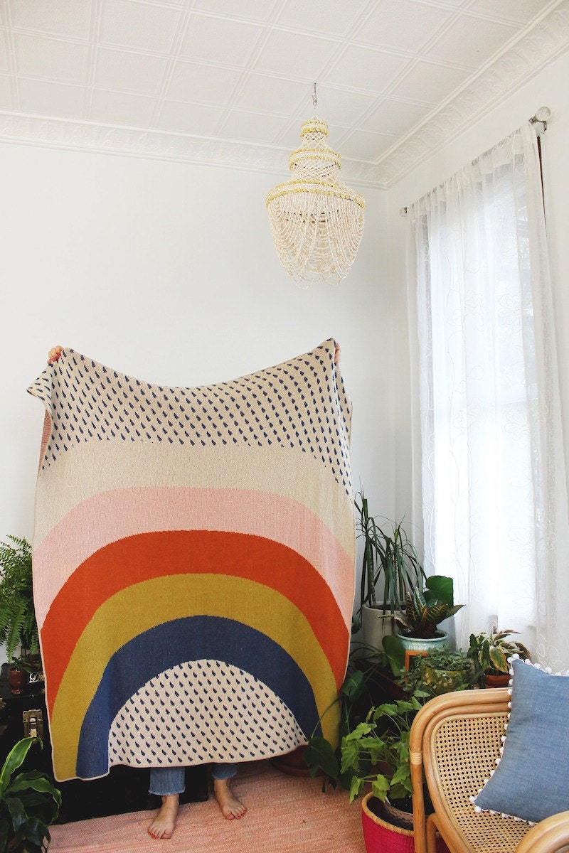 Person holds a rainbow knit blanket up in a white room filled with plants