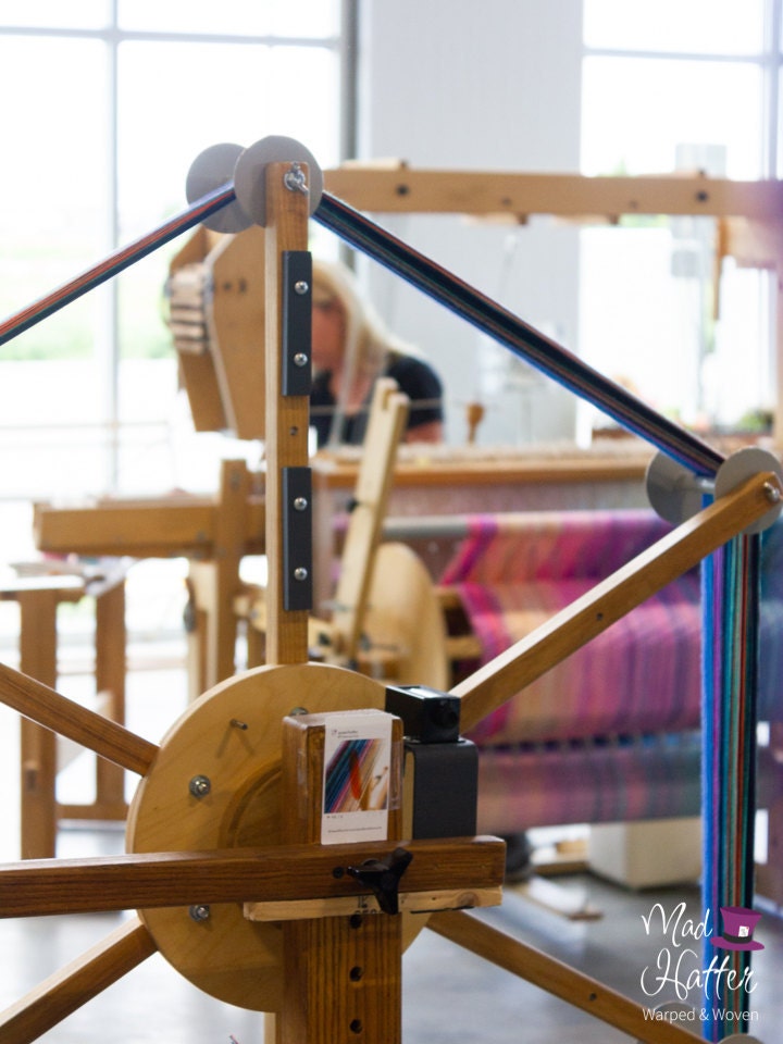 A wooden warping wheel inside of Mad Hatter Warped & Woven Studio. Wheel has blues, purples, greens, and other colours of thread on it. In the back, Wildflower is blurry and is weaving on a wooden loom with threads of pinks, creams, and blues. There are large windows with sunlight coming through in the background.