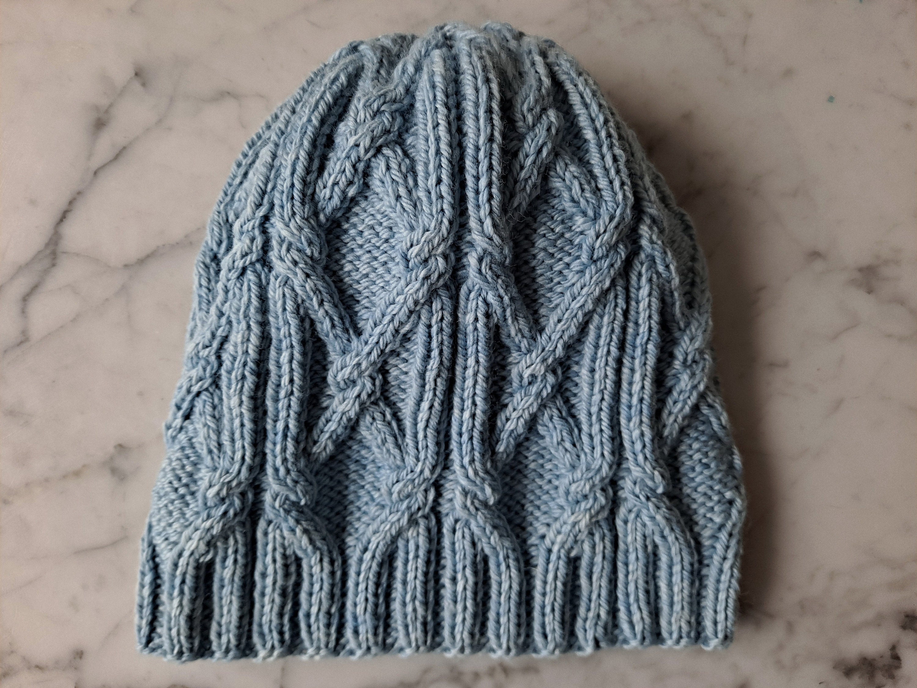 A pale blue cabled hat lies flat on a marble background.