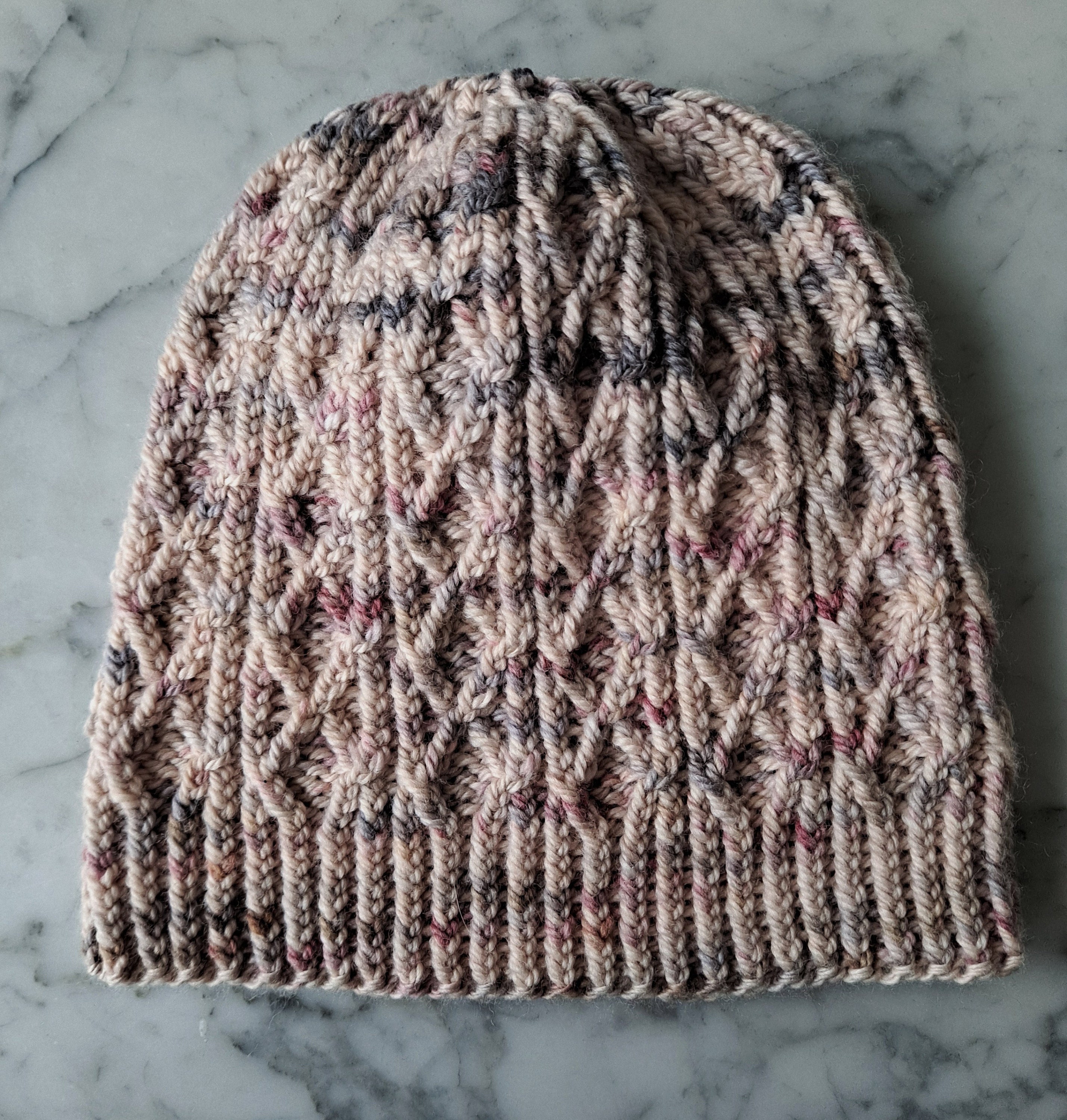 A pale pink cabled knitted hat lies flat on a marble background.