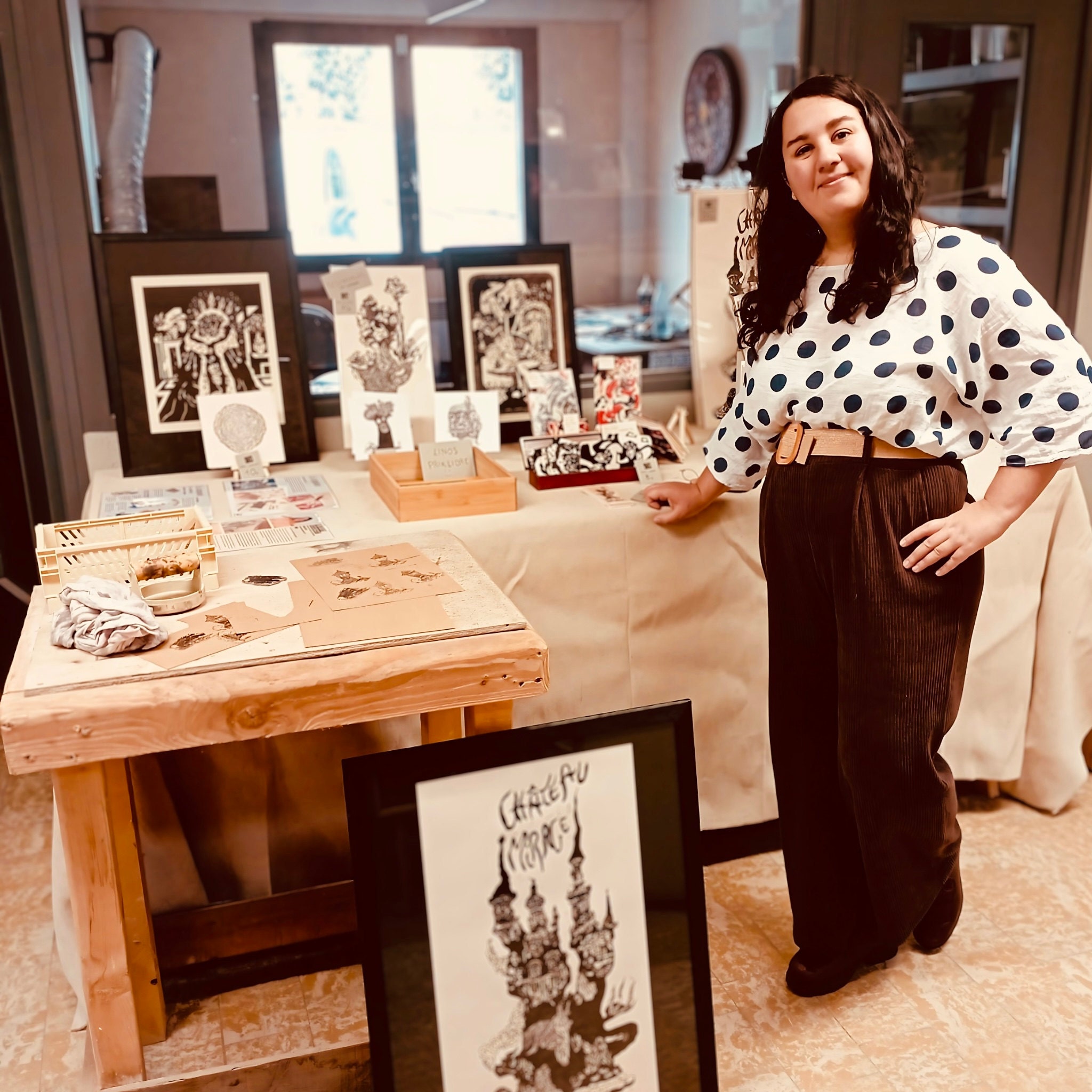 Portrait d’Hélène Bénistant devant son stand de créations en linogravure à Komuna. Elle se tient debout, souriante et en tenue élégante pour présenter ses œuvres artisanales. Les Linogravures sont présentées encadrées et bien ordonnée sur une table.