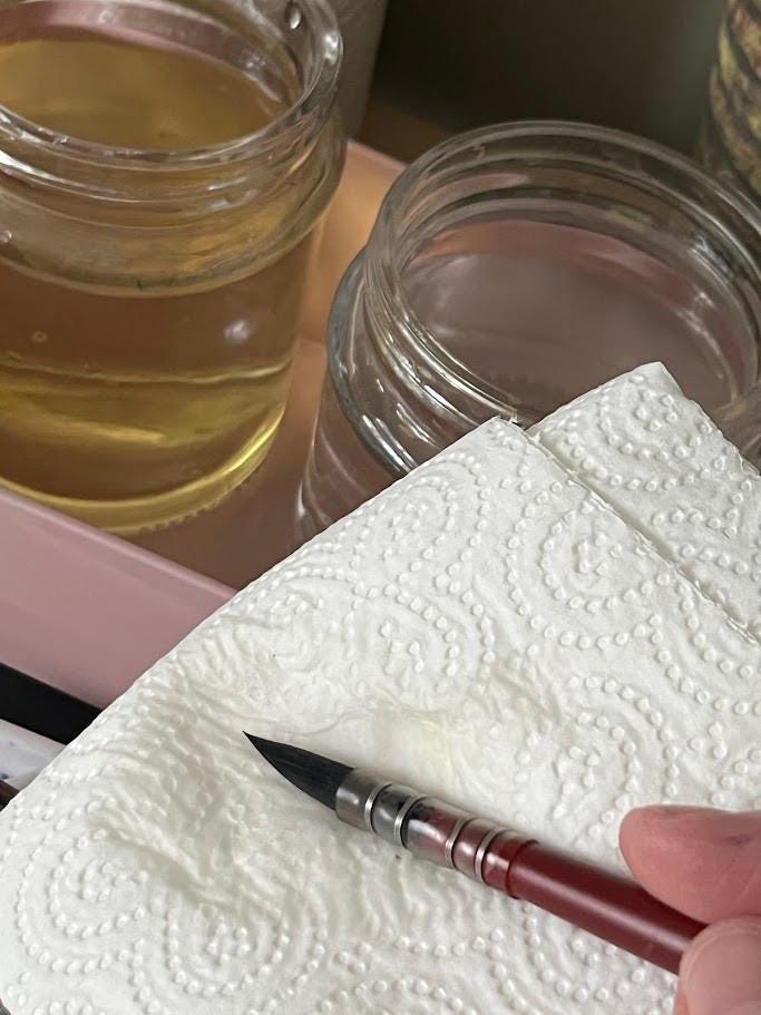 watercolour brush and water in pots, rinsed and being dabbed onto paper kitchen towel to remove excess water.