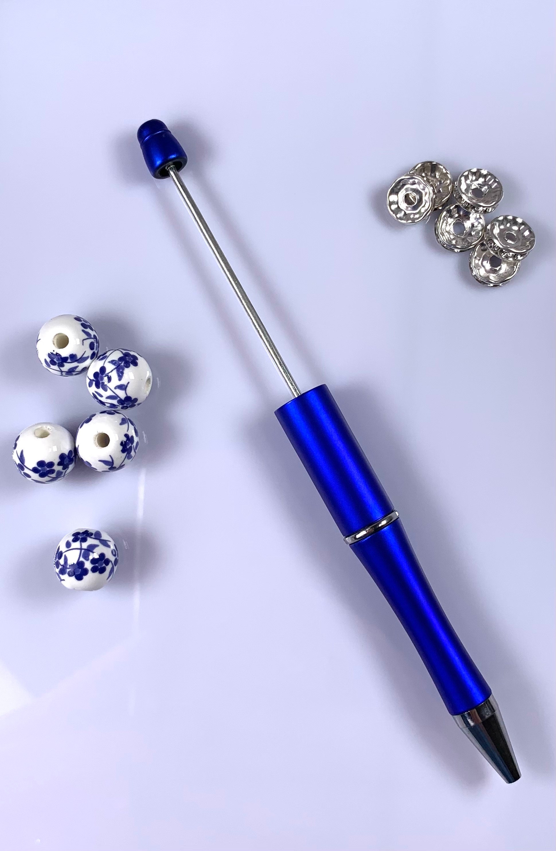 A vibrant blue beadable pen with a removable pin, lying on a white background. Nearby are five white ceramic beads decorated with blue floral patterns and several silver spacer beads, all ready for customization on the pens pin.