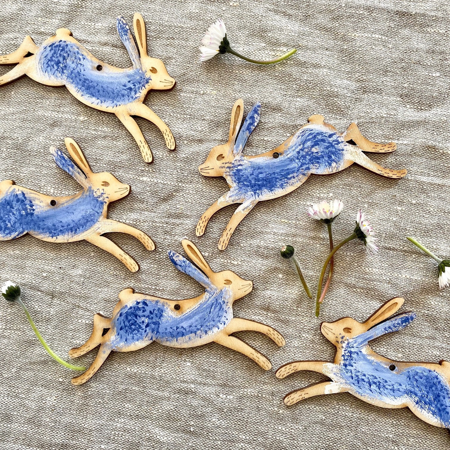 a picture of hand painted wooden hares ina blue and white colour palette on a natural linen background with daisies