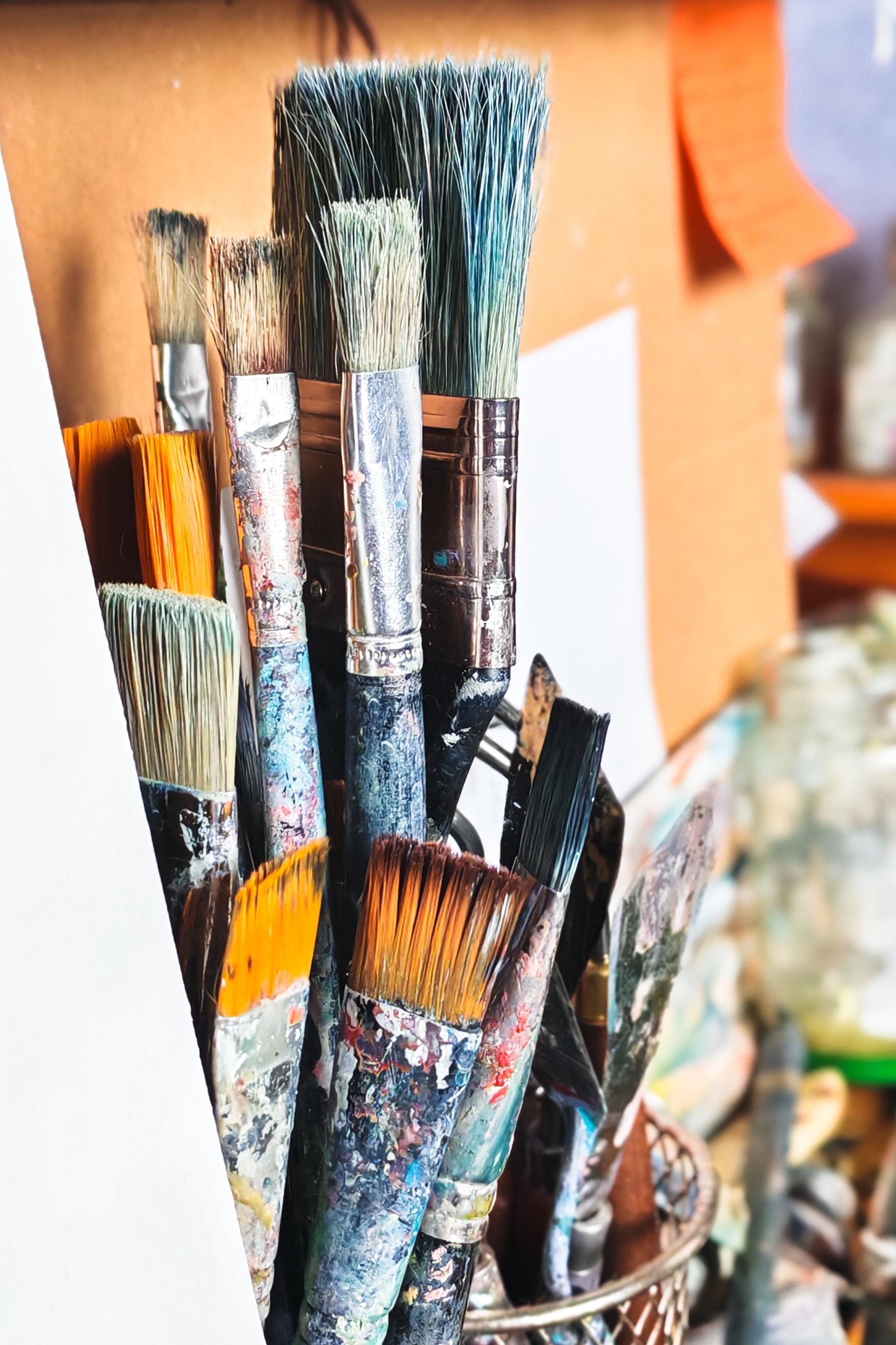 A collection of oil painters brushes, sitting upright beside a white canvas on a messy artists desk.