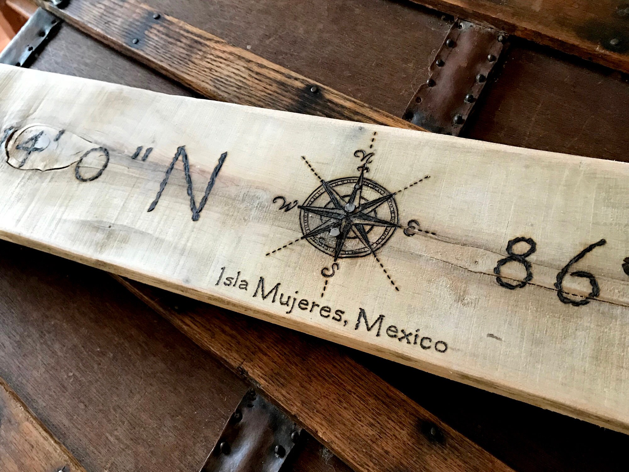 This picture shows a handmade location sign. It is a 36 inches long and 5 inches tall. The personalized coordinates are burned onto the wood. a decorative compass rose is in the center.