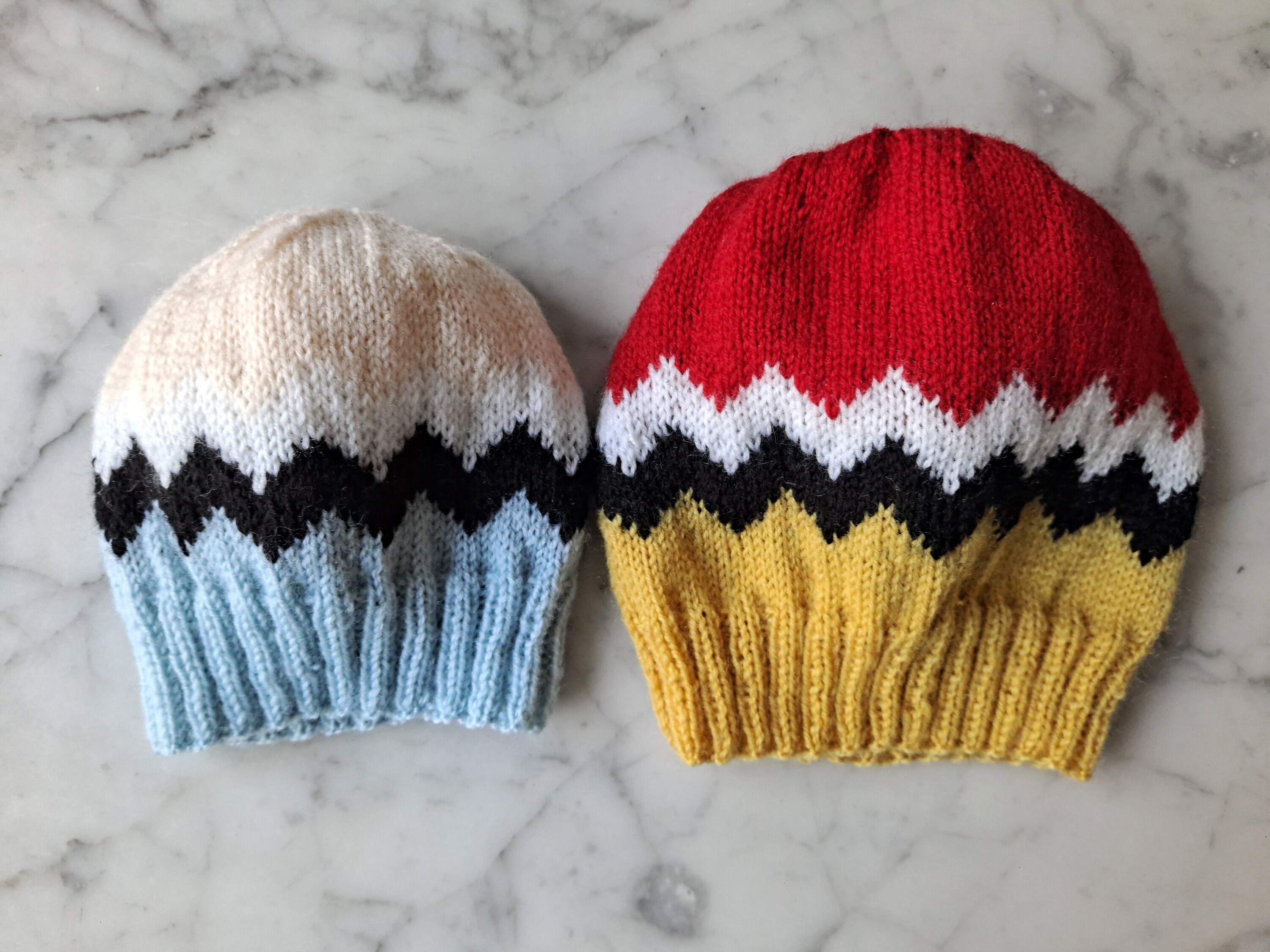 Two colourful beanie hats in different sizes lie side by side on a marble background. Both hats have bold black and white zig zag stripes, with different colours above and below the stripes.