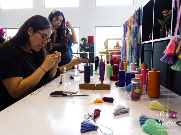 Guests are sitting at a Mad Hatter Warped & Woven work table making yarn tassels in all colours. There are spools of thread, tassels, scissors, and other supplies on the table. Guests look concentrated in their tassel making.