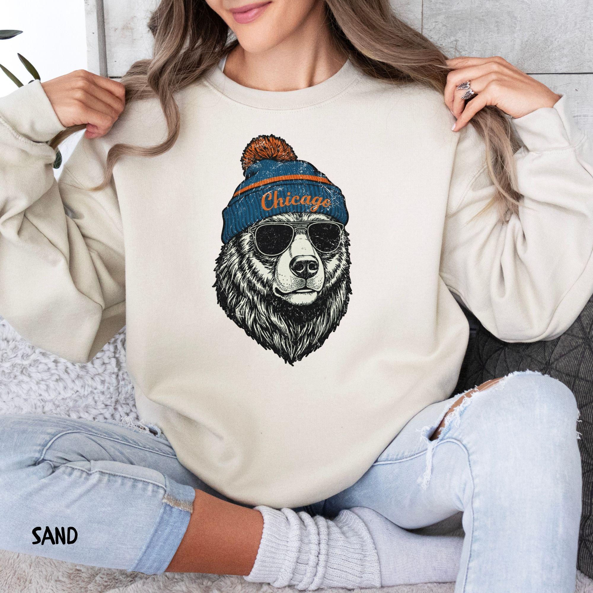 A woman wearing an ash gray sweatshirt featuring the vintage Chicago bear graphic while sitting comfortably on a couch.