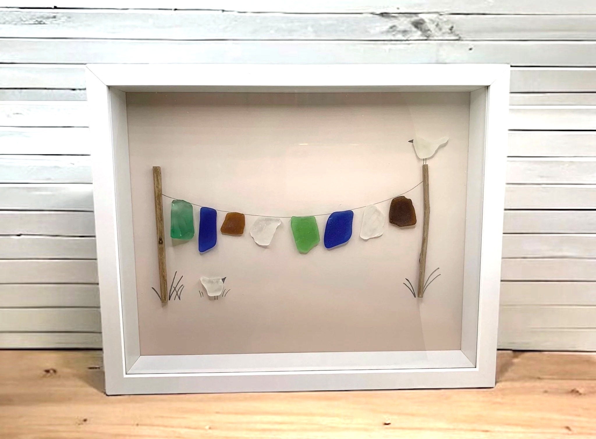 A framed piece of sea glass art featuring colorful sea glass pieces strung on a line between two wooden posts, resembling a whimsical outdoor display. The sea glass pieces include shades of green, blue, brown, and white. The design is enhanced with small drawn details, such as grass and a bird perched on one of the posts, set against a simple tan background and framed in a white shadow box frame.