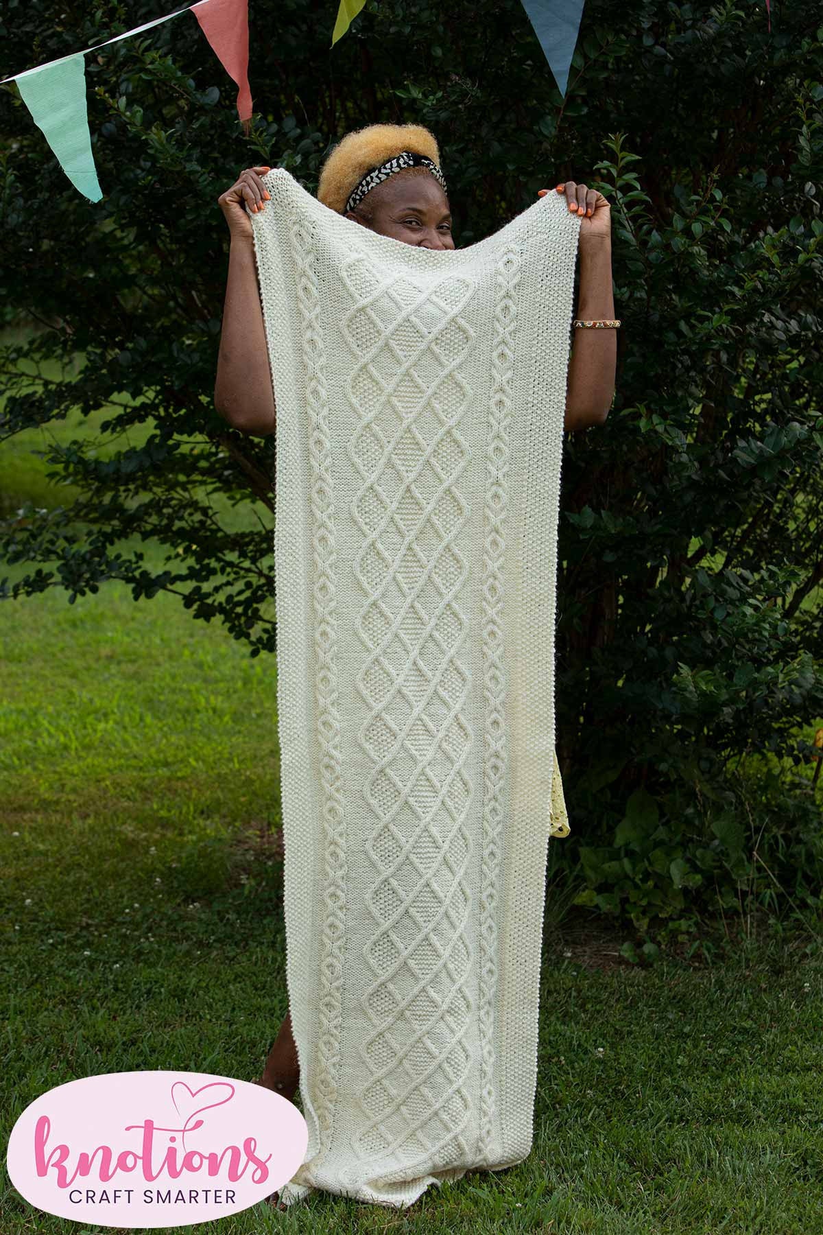 A smiling woman stands behind the shawl, which she is holding at one end to reveal the entire shawl. The shawl has a cabled Aran knitted pattern in white wool.