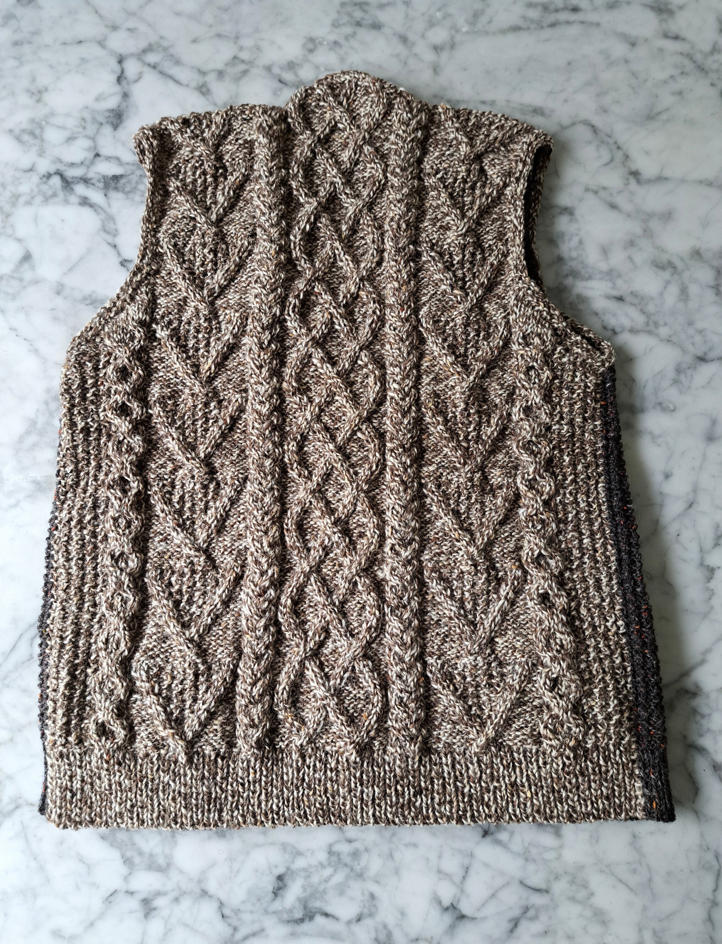 a light brown sleeveless Aran knit waistcoat lies flat on a marble background