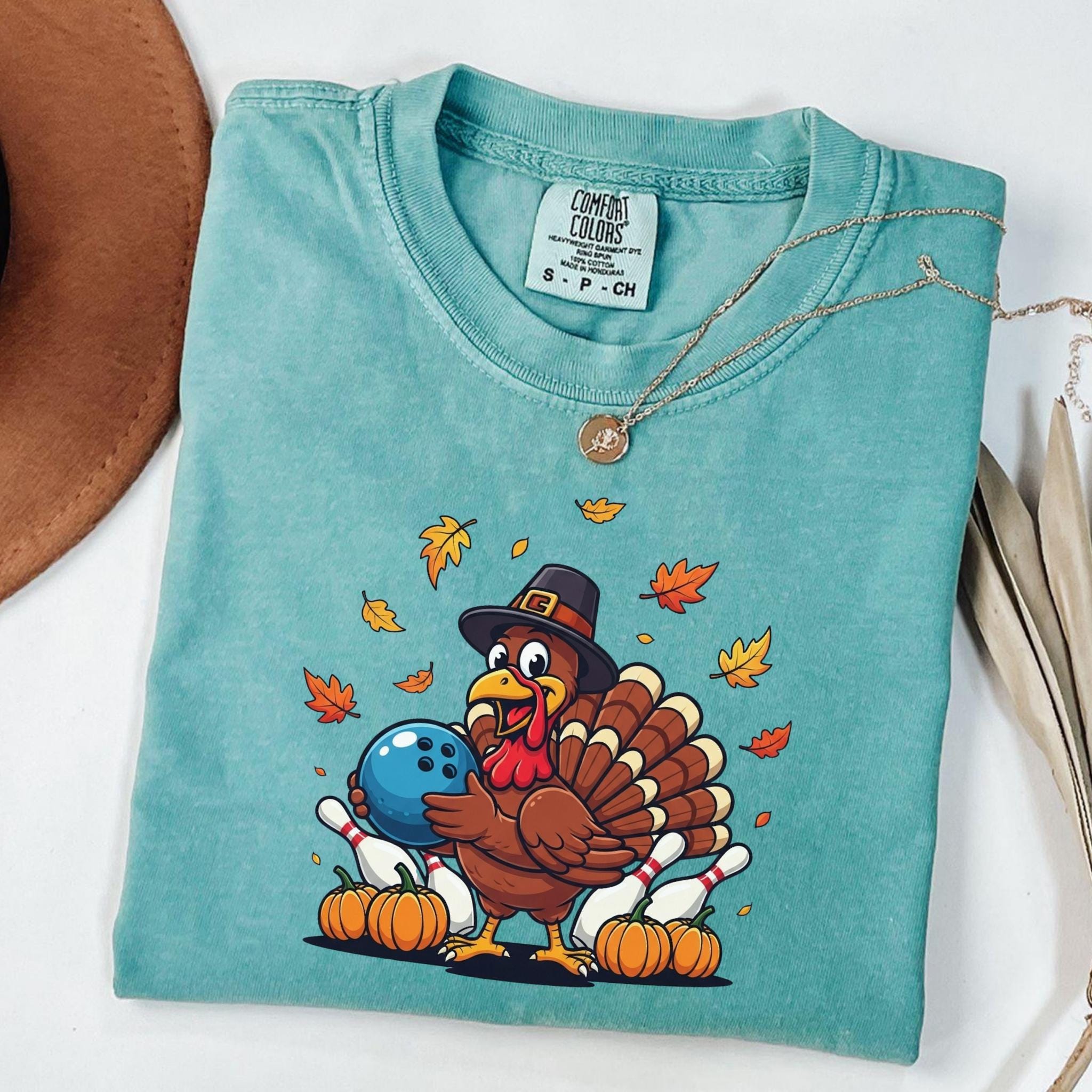 Funny cartoon turkey in a pilgrim hat holding a blue bowling ball surrounded by pumpkins, bowling pins, and autumn leaves. Cute Thanksgiving humor shirt for bowlers and fall lovers.