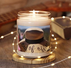 A luxuriously-scented candle featuring an image of a woman in a hat that reads out of office. The scented candle is lit.