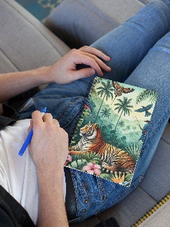 A person relaxing ready to begin to journal. They are holding a blue ballpoint pen and have a beautiful, custom-created spiral journal on their lap. The journal is decorated with a beautiful hand-created jungle image including a tiger.