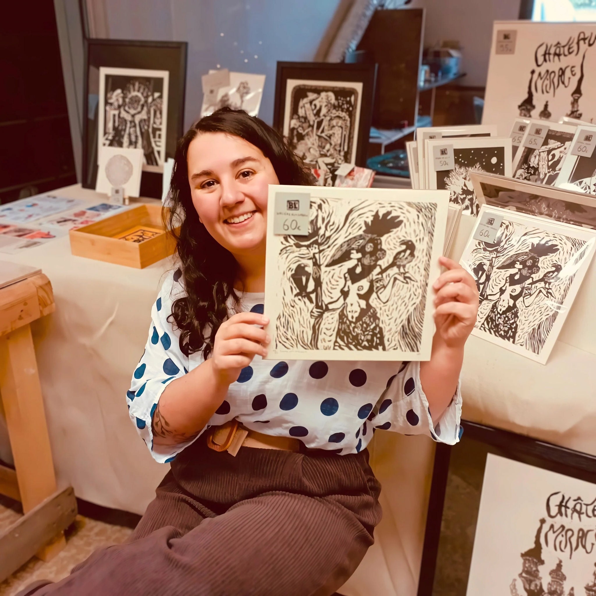 Portrait d’Hélène Bénistant, artiste linograveuse, souriante et élégante devant son stand d’exposition à Komuna. Elle tient une linogravure figurative représentant une sorcière et des corbeaux. Autour d’elle ses créations sont disposées sur des présentoirs, dans un cadre artisanal accueillant et festif.