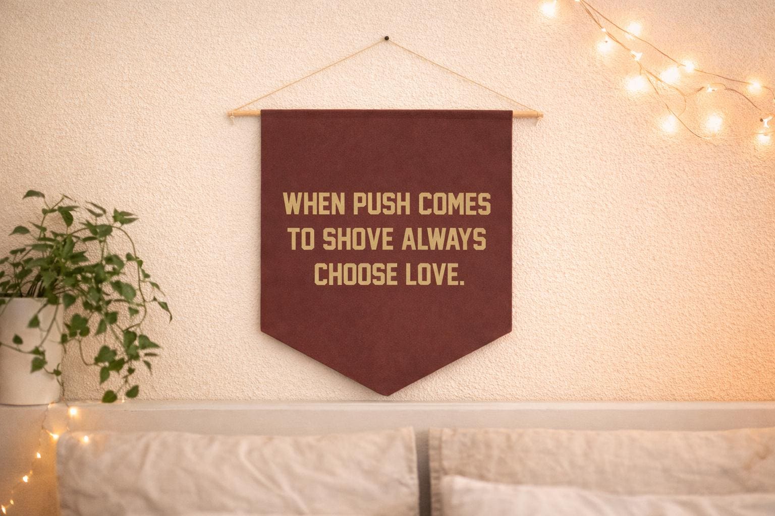 A dark burgundy felt pennant banner hanging on a light beige wall by a wooden dowel with string. The banner reads WHEN PUSH COMES TO SHOVE ALWAYS CHOOSE LOVE. in bold gold lettering. A trailing green pothos plant in a white pot sits to the lower left, and warm string lights decorate the upper right corner and shelf below. A light gray sofa is partially visible at the bottom of the frame.