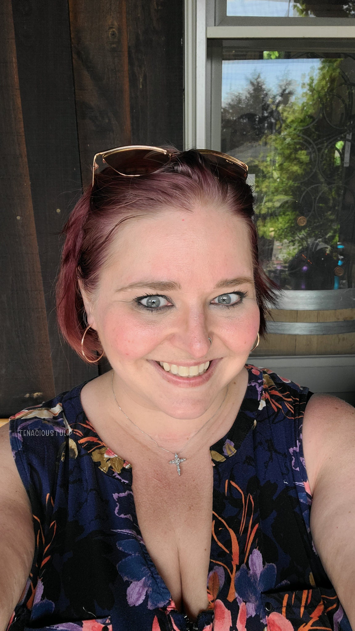 Woman with red hair and sunglasses on her head. She is wearing a navy blue floral sleeveless dress taking a selfie.
