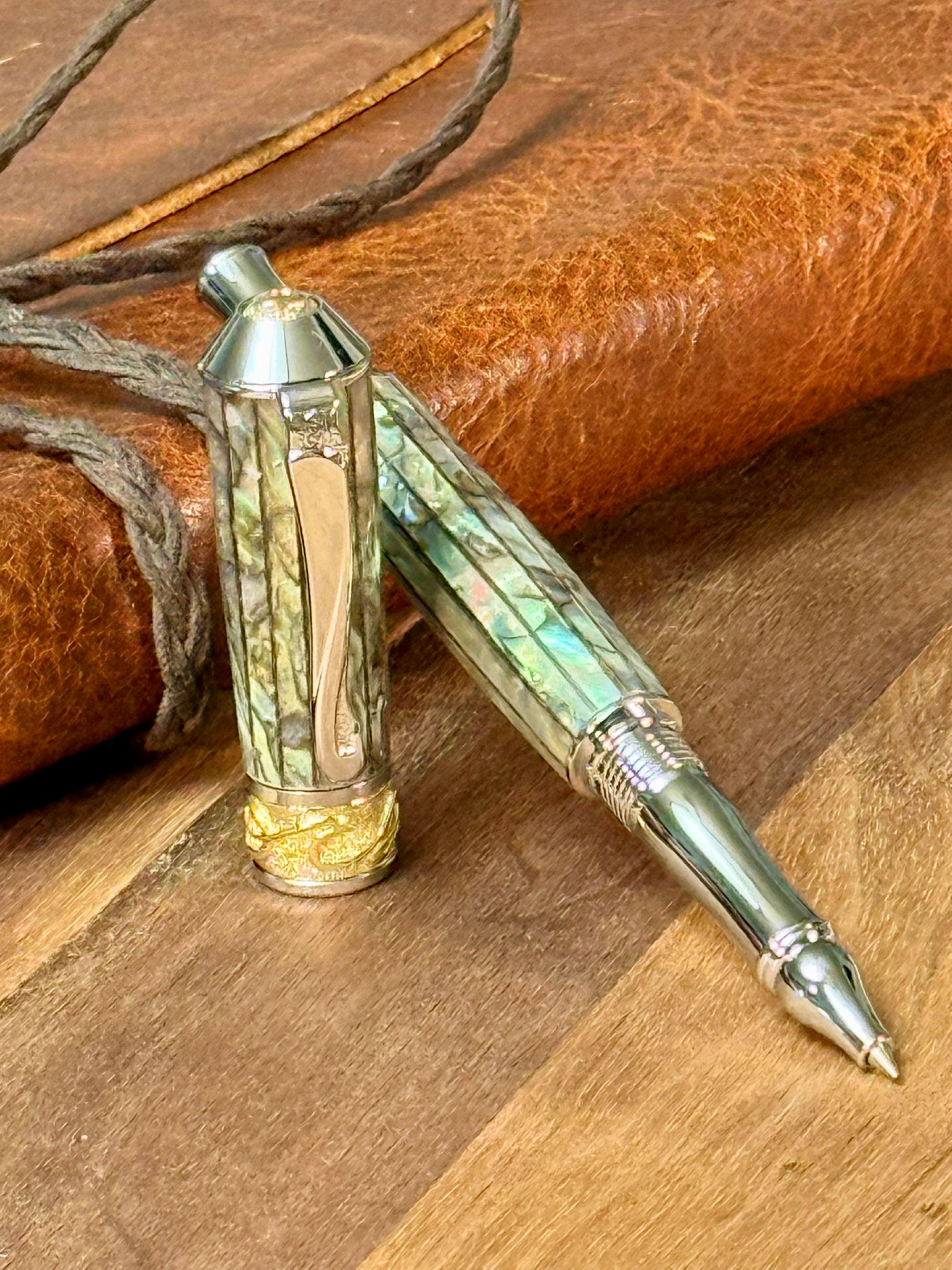 The Nouveau Sceptre Rollerball pen shown with Paua Abalone Body, Platinum and 22k Gold Trim. The pen is leaning against a leather journal on wooden table.