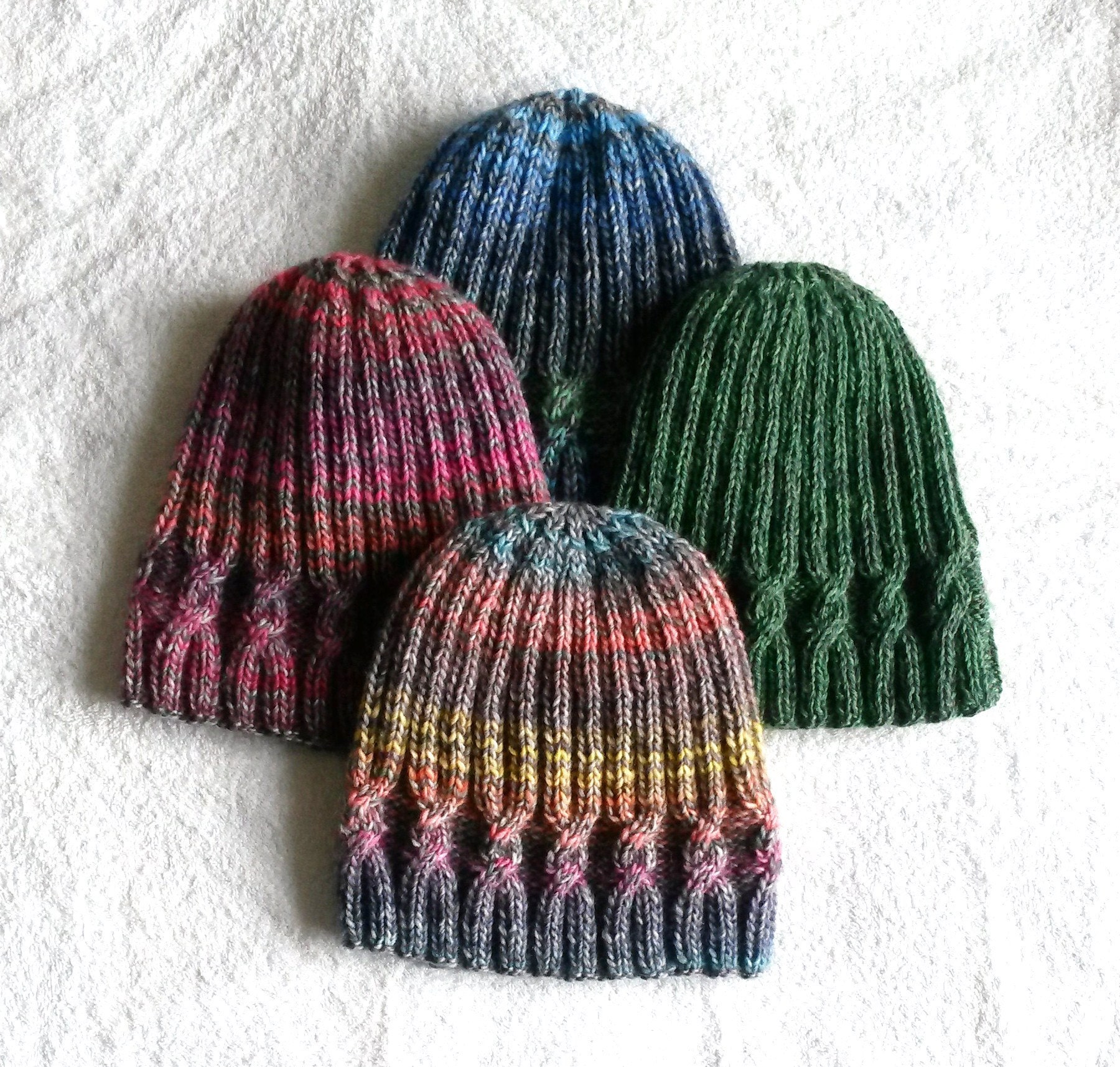 Four Simple Cable Beanie hats of different colours lie flat on a white background.