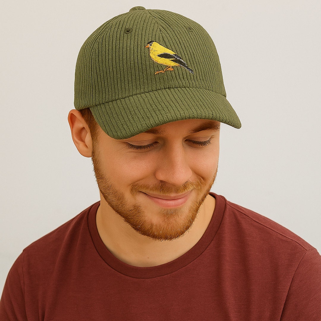A man in a maroon t-shirt wears an olive green corduroy baseball cap embroidered with a yellow goldfinch. He smiles gently with his eyes closed, standing against a light, neutral backdrop.