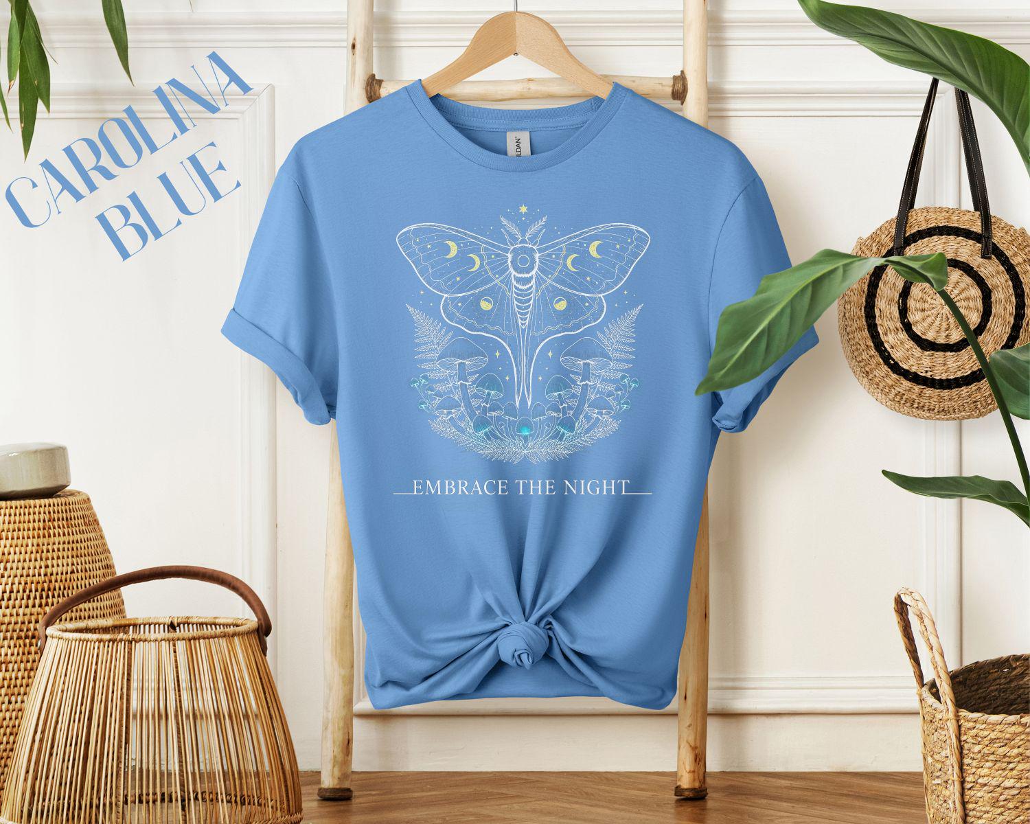 Black unisex T-shirt on hanger featuring celestial moth, moons, and mushrooms with ‘EMBRACE THE NIGHT’ text in a cozy home setting.
