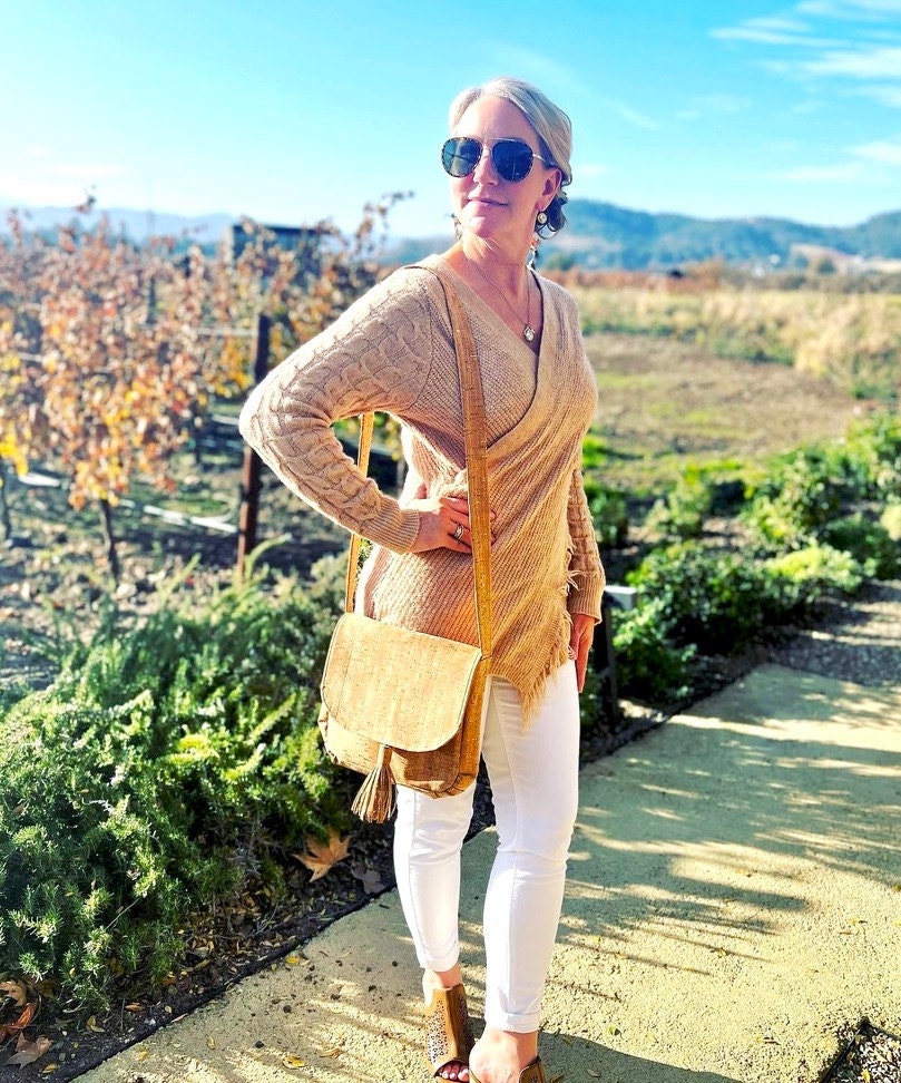 Woman in vineyard wearing white jeans, an camel sweater and booties, with a cork purse