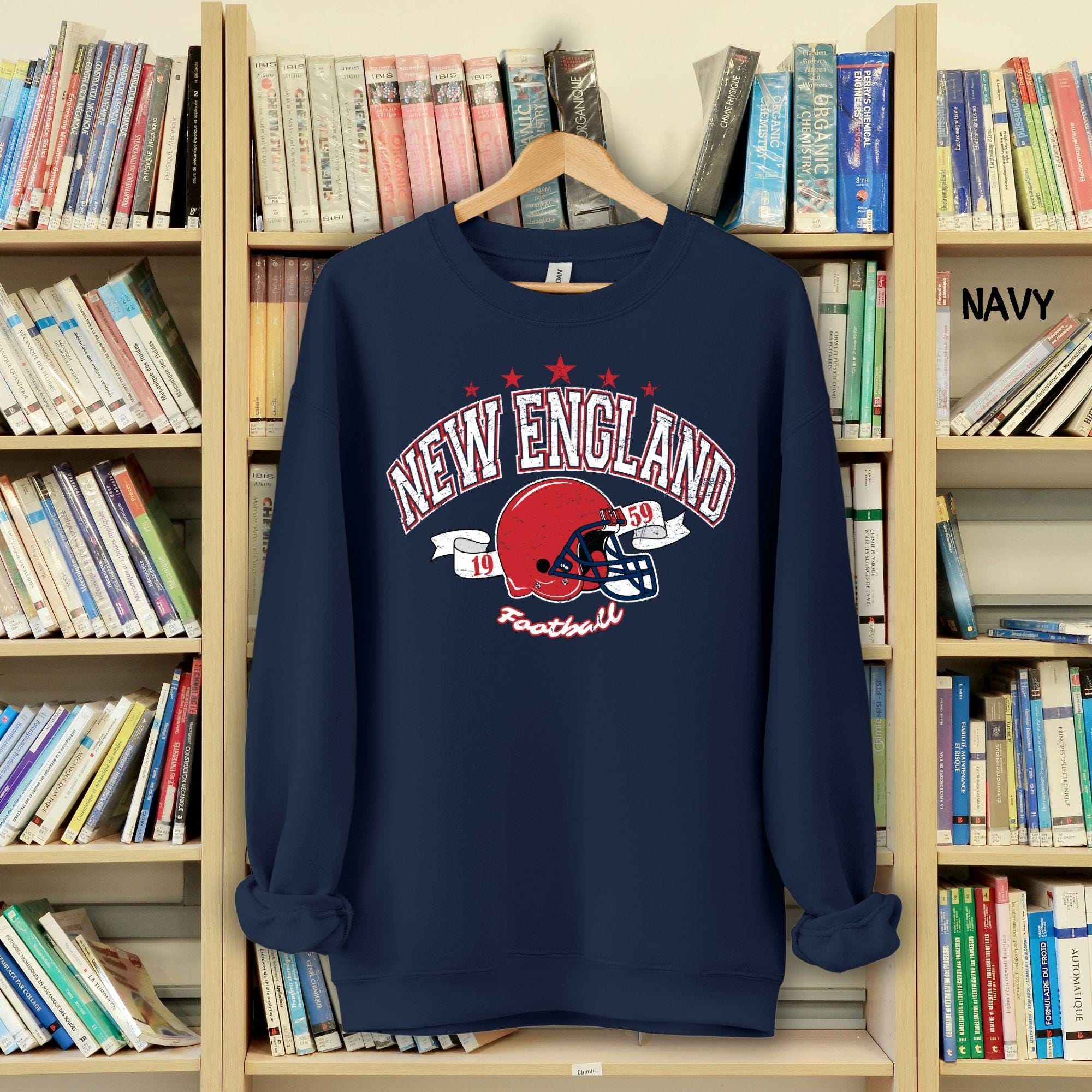 A person wearing a vintage-style navy blue crewneck sweatshirt with a distressed New England Football helmet design on a bookshelf background.