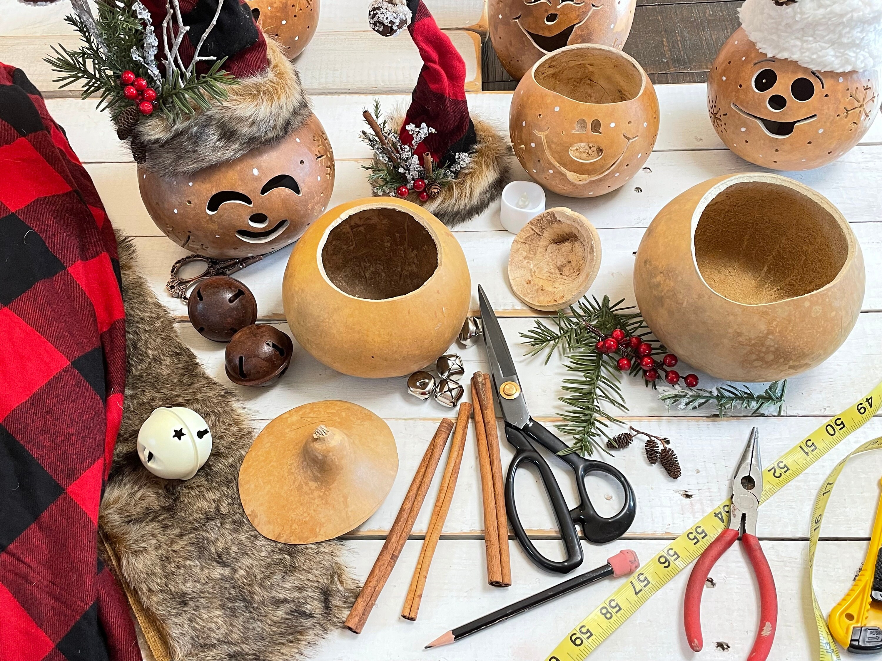 A collections of dried gourds cut and cleaned with their tops off. Buffalos check fabric, faux fir, cinnamon sticks, and jingle bells are laid out to use for the handmade Christmas hats.