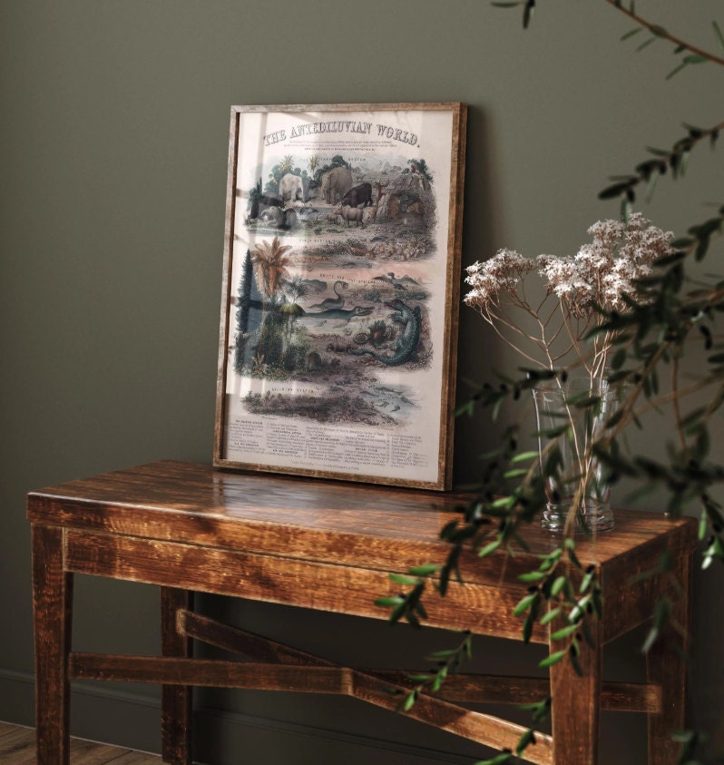 A framed vintage lithograph titled The Antediluvian World resting on a rustic wooden table with a vase of dried white flowers, set against an olive-green wall, showcasing intricate illustrations of prehistoric life and geological eras