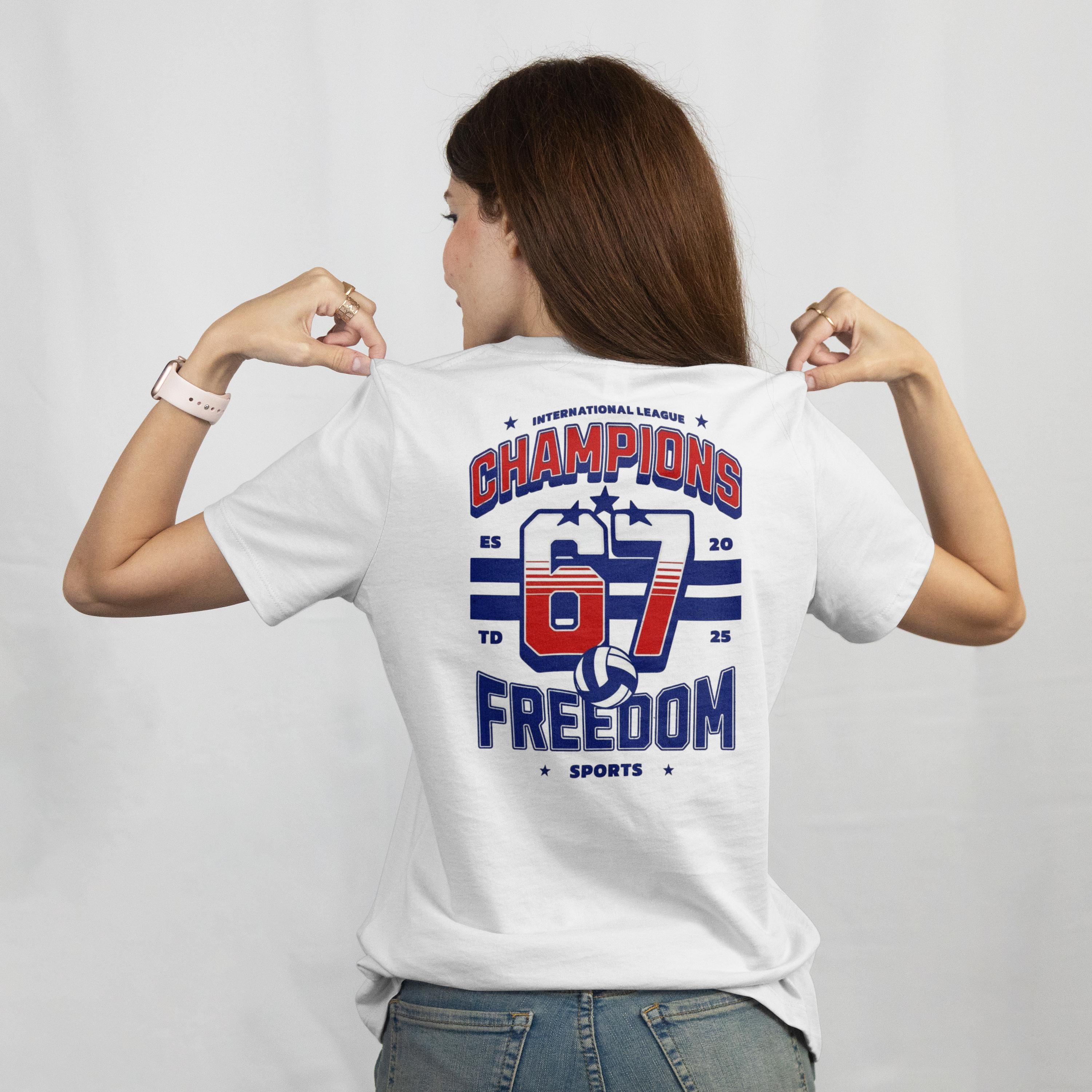 back view of a woman wearing a white tshirt with bold red and blue graphic text that reads Champions 67 Freedom Sports. She is pointing to the design on her back with both thumbs. The setting is plain, neutral background.