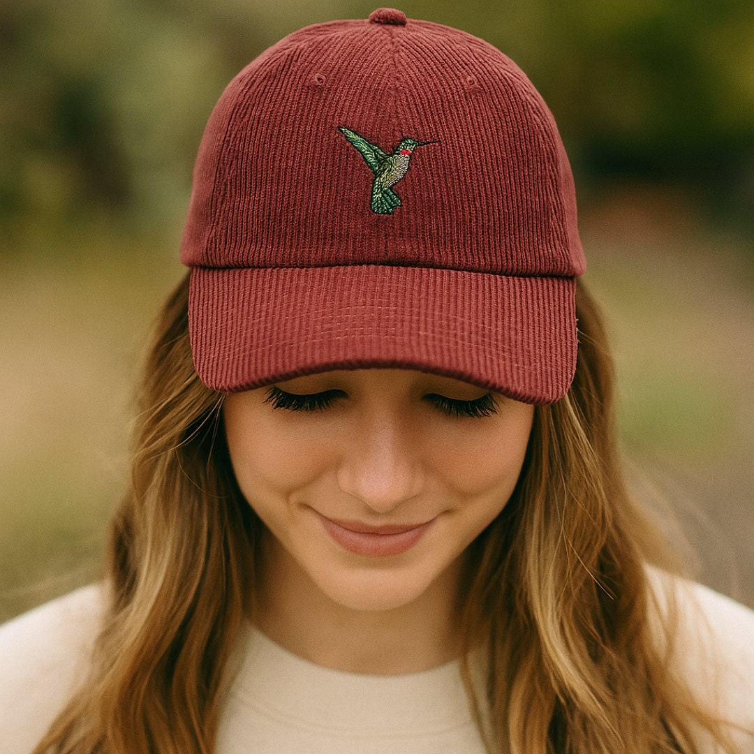 A young woman wearing a burgundy corduroy baseball cap embroidered with a green and red hummingbird. She smiles gently while standing outdoors with a soft, natural background.