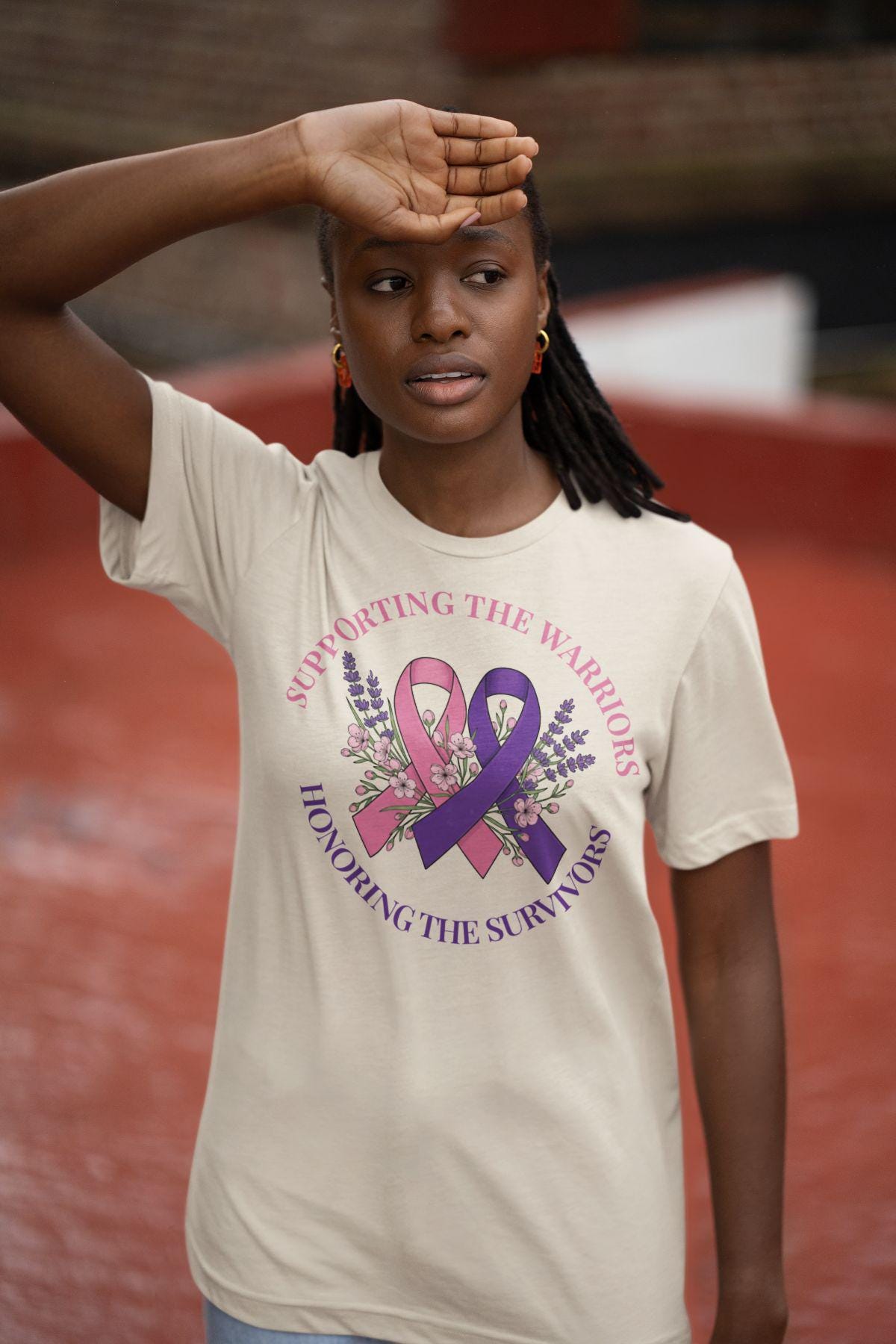 Woman with braided hair looking thoughtfully to the side while wearing a natural-colored awareness shirt with a pink and purple ribbon design.