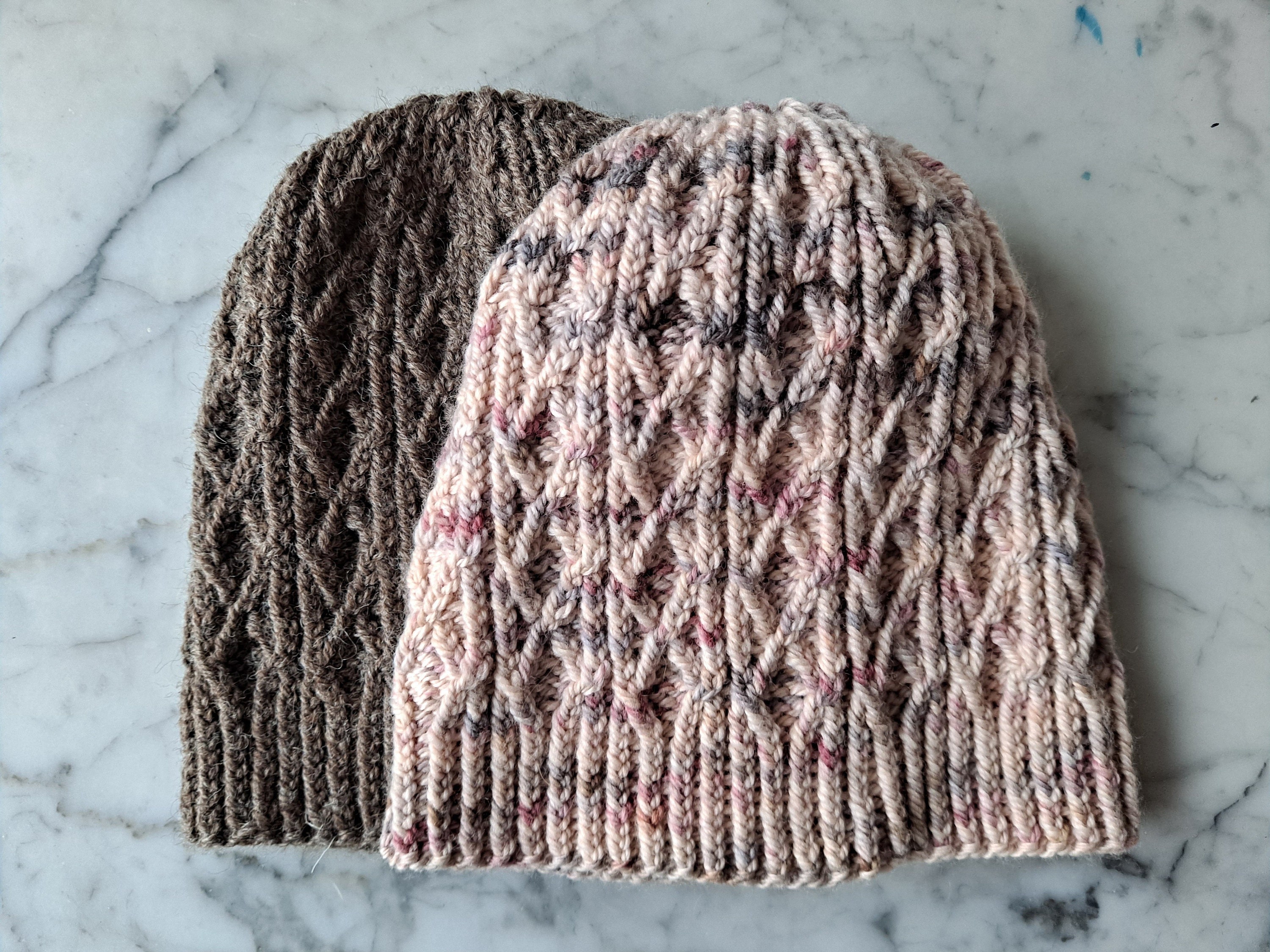 Burren Beanie: two hats lie flat on a marble background. Both feature a cabled lattice pattern.