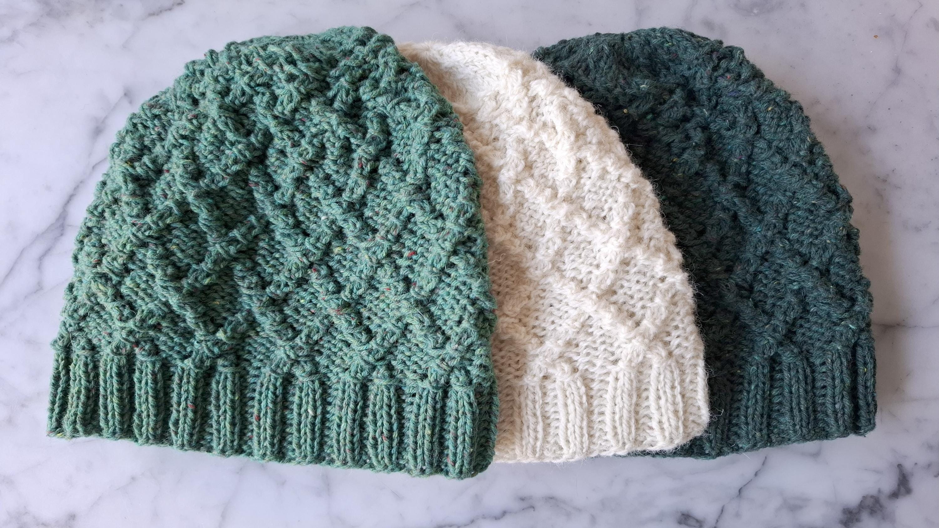 Three handknit cabled hats in shades of green and white lie flat on a marble background