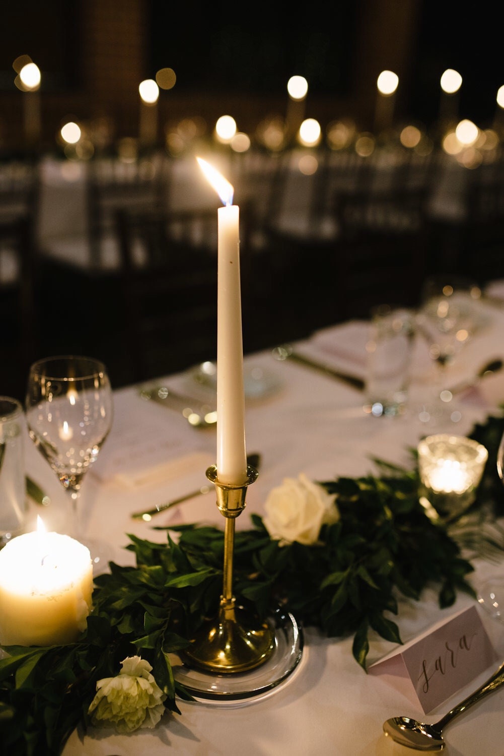 Photo of wedding reception table decorated with gold calligraphy placecard, green foliage and long candles