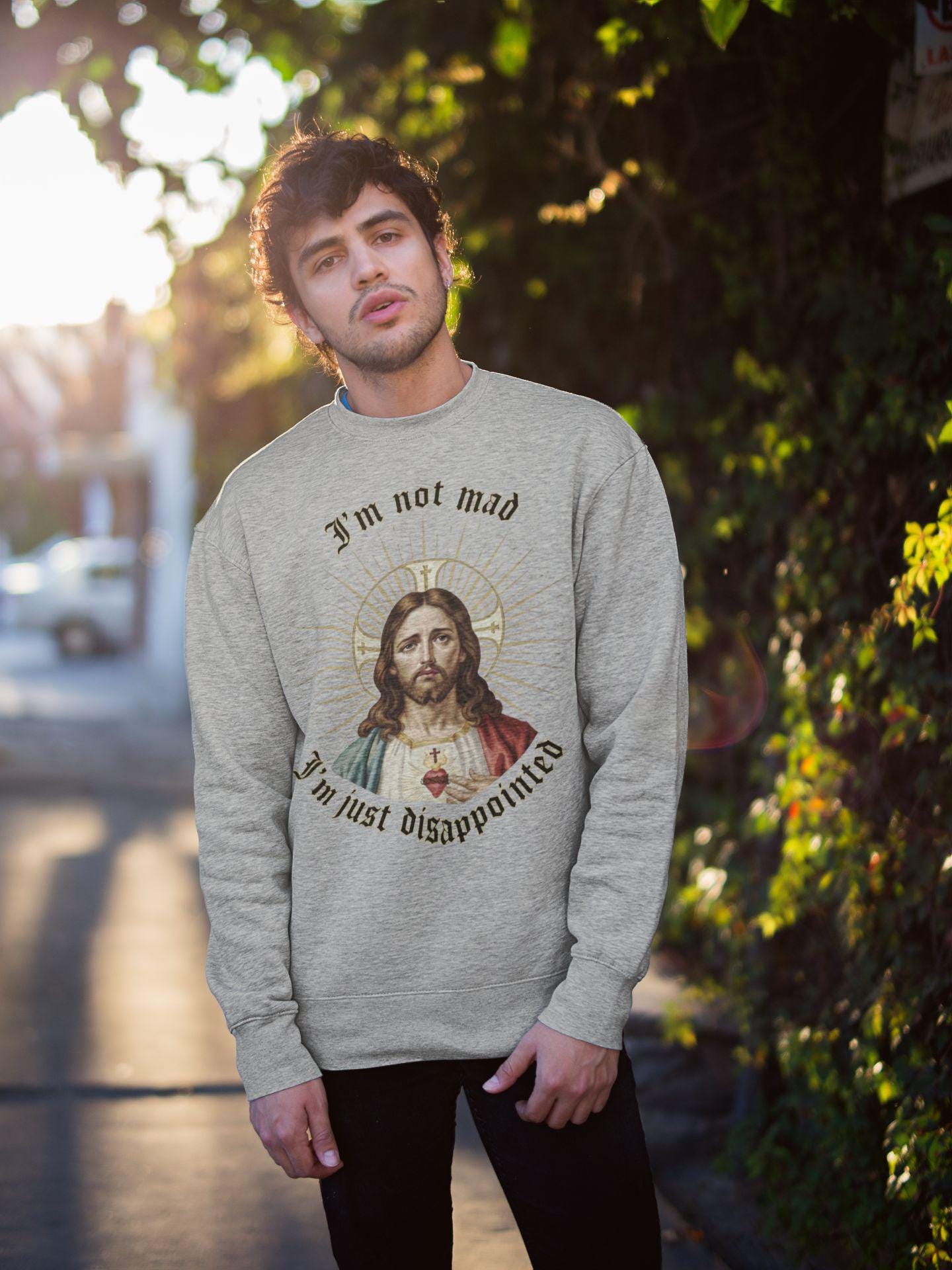 A stylish man in an urban setting wearing a white sweatshirt. The sweatshirt features a funny meme graphic of Jesus with the text Im not mad, Im just disappointed.