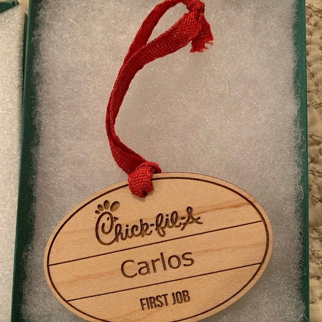 A solid maple hardwood chick-fil-a name tag, turned into an hanging tree ornament, with a red ribbon running from the top to hang it by. It is engraved with Carlos and First Job below.
